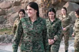 L'image montre un groupe de femmes en uniforme militaire, marchant ensemble dans un environnement extérieur. L'une d'entre elles, qui se trouve à l'avant, sourit et semble confiante. Les autres femmes l'accompagnent, toutes portant des uniformes camouflés. Le décor autour d'elles semble naturel, avec des rochers et de la végétation. L'atmosphère générale est positive et enjouée.