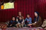 L'image montre une mère assise avec quatre enfants dans une pièce décorée de manière traditionnelle. La mère porte un vêtement coloré et un foulard, tandis que les enfants, d'âges variés, sont assis autour d'elle. L'un des enfants est plus jeune et semble dans les bras d'un autre enfant. L'arrière-plan est orné de tissus aux motifs riches, et il y a une lumière douce qui entre par la fenêtre, créant une atmosphère chaleureuse et conviviale.