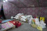 L'image montre l'intérieur d'une tente décorée avec un motif coloré sur les murs. À l'intérieur, on peut voir des tas de sacs et de paquets de différentes tailles, probablement des produits alimentaires ou de la marchandise. Ces sacs sont empilés sur des palettes en bois. Dans un coin, il y a des couvertures ou des tapis. L'éclairage est doux, créant une ambiance chaleureuse dans cet espace.