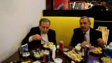 L'image montre deux hommes assis à une table dans un restaurant. L'un d'eux est en train de manger avec une cuillère, tandis que l'autre semble discuter. Les deux hommes portent des costumes et sont entourés de nourriture, y compris des assiettes pleines et des canettes de boissons. L'ambiance semble décontractée, avec un décor moderne en arrière-plan.