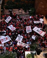 L'image montre une grande foule rassemblée dans un espace public, tenant des pancartes avec des slogans revendiquant des droits et faisant des déclarations politiques. La majorité des personnes portent des vêtements rouges. L'ambiance semble mobilisée et énergique, avec des palmiers décoratifs autour.