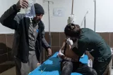 در این تصویر، یک بیمار در یک تخت بیمارستانی در حال درمان است. یک پرستار در حال پانسمان جراحت بیمار در ناحیه سینه اوست. مردی دیگر که لباس معمولی به تن دارد، در حال نگه داشتن یک بطری آب یا سرم است. فضای اتاق بیمارستان ساده و کارکردی به نظر می‌رسد. دیوارها دارای رنگ ملایم و تخت بیمارستان آبی است. این تصویر نشان‌دهنده تلاش کادر پزشکی برای ارائه مراقبت به یک بیمار است.