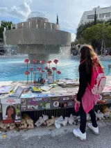 L'image montre une jeune fille devant une fontaine. Elle semble observer un mémorial ou un hommage qui se trouve sur le bord de la fontaine, où sont disposés des photos, des fleurs et divers objets en mémoire d'une personne. Il y a aussi des peluches blanches sur le sol, ce qui ajoute une touche émotive à la scène. La fontaine est entourée de verdure, ce qui donne une atmosphère paisible malgré la gravité de l'hommage. La fille, portant un sac à dos, semble pensive.