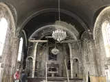 L'image montre l'intérieur d'une église en ruine. Le plafond est voûté et présente des traces de délabrement, avec des murs en pierre grise exposant des signes d'usure et de dégradation. Un lustre pend au centre, bien qu'il semble en mauvais état. Deux personnes se tiennent au centre, probablement en train d'évaluer l'endroit. Des échafaudages sont visibles, suggérant des travaux de restauration ou de réparation. L'atmosphère est marquée par un sentiment de désolation mais aussi de potentiel renouveau.