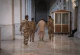 L'image montre quatre hommes marchant ensemble dans un couloir large et éclairé. Ils portent des uniformes militaires et semblent se diriger vers une sortie. Le sol est en marbre, et les murs sont ornés, créant une ambiance à la fois solennelle et majestueuse.