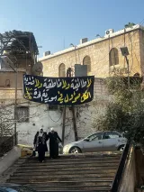 L'image montre deux femmes en hijab marchant dans une rue avec une maison à l'arrière-plan. Un grand panneau noir et jaune est accroché, portant un message en arabe. Le ciel est dégagé, et des élements architecturaux rustiques sont visibles.