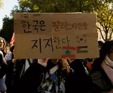 L'image montre une personne tenant une pancarte en carton lors d'une manifestation. La pancarte est écrite en coréen et affiche un message de soutien aux Palestiniens. En bas, il y a également des drapeaux représentant le Liban et la Palestine. L'arrière-plan montre une foule de manifestants, entourée d'arbres, suggérant que l'événement se déroule en plein air.