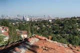 L'image montre un panorama d'une ville, avec une vue depuis une colline. Au premier plan, on peut voir des toits en tuiles rouges, tandis qu'à l'arrière-plan, une vaste étendue de bâtiments modernes et de gratte-ciel se dresse contre un ciel bleu clair. La végétation est également présente, avec de nombreux arbres dispersés sur les collines, créant un contraste entre la nature et l'urbanisation. L'ensemble donne une impression de calme et de vastitude, mettant en valeur la coexistence entre les zones urbaines et rurales.