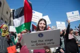 L'image montre une manifestation avec des participants brandissant des pancartes et des drapeaux. Au premier plan, une femme tient une pancarte avec le message "End the siege on Gaza strip" (Mettre fin au siège de la bande de Gaza) tout en levant un drapeau palestinien. Les manifestants semblent exprimer des préoccupations concernant la situation à Gaza, en particulier en ce qui concerne les femmes et les enfants. L'ambiance est déterminée et solidaire.