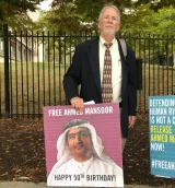 L'image montre un homme debout, habillé en costume avec une cravate, tenant une grande pancarte. La pancarte présente un portrait d'un homme arabe, probablement Ahmed Mansoor, avec un message qui célèbre un anniversaire et demande sa libération. Le fond de l'image est constitué d'un paysage urbain, avec des arbres et une clôture derrière lui. L'homme semble participer à une manifestation ou un rassemblement pour défendre les droits humains.