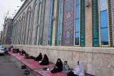 L'image montre un mur orné de mosaïques colorées, typiquement islamique, avec de grandes fenêtres. Au premier plan, on voit plusieurs femmes assises, portant des vêtements traditionnels, sur des tapis ou des morceaux de tissu au sol. Elles semblent discuter ou prier. La scène dégage une atmosphère paisible, et on peut apprécier la beauté architecturale du lieu.