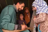 L'image montre un homme assis, en train d'écrire sur un support en bois, avec un crayon à la main. Autour de lui, plusieurs jeunes filles l'observent attentivement, certaines portant des foulards. L'environnement semble être une pièce ou un espace informel, peut-être une tente, avec des motifs colorés ou traditionnels. L'atmosphère est concentrée et engageante, suggérant un moment d'apprentissage ou d'enseignement.
