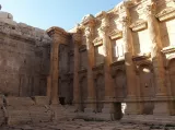 L'image montre un ancien bâtiment en ruines, probablement un temple ou un théâtre, avec des colonnes majestueuses et des murs en pierre. La lumière du soleil éclaire l'intérieur, mettant en valeur les détails architecturaux et les textures du matériau. Des marches descendent vers le sol, et on peut observer des vestiges de la construction. L'ambiance de l'image évoque l'histoire et le caractère ancien du site.