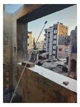 L'image montre une vue à travers une ouverture dans un mur, probablement d'un bâtiment abandonné ou en ruines. Au premier plan, on remarque un long objet métallique posé sur le rebord de la fenêtre. La scène révèle des bâtiments voisins, certains intacts, d'autres en mauvais état, avec des signes visibles de dégradation. Les murs sont couverts de graffitis et d'écritures, ajoutant une touche de couleur et d'expression à l'environnement urbain. La lumière du matin éclaire la scène, créant une atmosphère mélancolique.