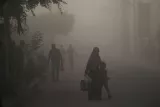 L'image montre une rue floue et brumeuse, probablement en raison de la pollution ou d'une tempête de poussière. On y voit plusieurs personnes marchant, dont une femme en hijab qui tient la main d'un jeune garçon. Ils portent des sacs. Les silhouettes des autres passants sont également visibles dans le brouillard, créant une atmosphère sombre et mystérieuse. Les arbres et les bâtiments en arrière-plan sont partiellement dissimulés par la brume.