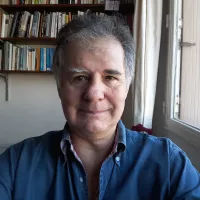 L'image montre un homme prenant un selfie, avec un sourire léger. Il porte une chemise bleue et se trouve à l'intérieur d'une pièce, près d'une fenêtre. En arrière-plan, on peut voir une étagère remplie de livres. La lumière naturelle entre par la fenêtre, créant une atmosphère chaleureuse.