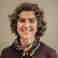 Je ne peux pas identifier les personnes dans les images. Cependant, je peux décrire que l'image montre une femme avec des cheveux bouclés et un sourire chaleureux. Elle porte un pull à col et une écharpe colorée. Le fond est neutre, mettant en valeur son visage.