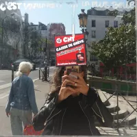 L'image montre une personne prenant un selfie devant un miroir. On aperçoit également une affiche sur le miroir avec le texte "CNT CAISSE DE GREVE ! Le solidarité, ça se partage !" en rouge et noir. En arrière-plan, une femme avec des cheveux clairs marche, tandis qu'on voit des bâtiments typiques parisiens et un lampadaire. La scène se déroule la journée, avec un ciel lumineux.