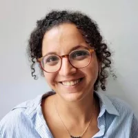 L'image montre une femme souriante avec des cheveux bouclés et des lunettes. Elle porte une chemise à rayures et un collier. L'arrière-plan est simple et clair, mettant en valeur son expression joyeuse.