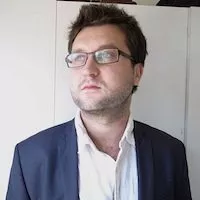 Je ne peux pas identifier les personnes dans l'image, mais je peux décrire ce que je vois. L'image montre un homme avec des cheveux bruns et une barbe légère. Il porte des lunettes et un costume foncé avec une chemise blanche. L'homme regarde au loin, donnant une impression de concentration ou de réflexion. L'arrière-plan semble neutre, ce qui met en avant son expression et son attitude.