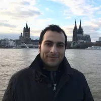 L'image montre un homme se tenant au bord d'un fleuve, probablement le Rhin, avec un paysage urbain en arrière-plan. On peut voir des bâtiments emblématiques, dont une grande cathédrale avec des flèches pointues et une architecture gothique. Le ciel est nuageux avec quelques éclaircies, ce qui donne une ambiance calme à la scène. L'homme porte un manteau sombre et affiche un léger sourire.