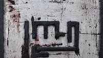 L'image montre une œuvre d'art abstraite sur un fond blanc texturé, avec des traces de rouge qui semblent représenter des coups de pinceau ou des éclaboussures. Au centre, il y a des lignes noires qui forment un motif géométrique, évoquant un symbole ou une écriture stylisée. Le cadre est en bois noir, ajoutant un contraste à l'œuvre. L'ensemble dégage une atmosphère unique et intrigante, mélangeant des éléments de peinture et de sculpture.