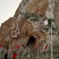 La imagen muestra un paisaje montañoso con una gran roca o acantilado en el centro, que tiene una cueva visible en su base. A su alrededor, hay varias banderas ondeando, entre las cuales algunas son de Argelia, ya que tienen un diseño distintivo. En la parte inferior de la imagen, se pueden ver algunas personas caminando y disfrutando del lugar. El cielo tiene un tono claro, posiblemente al atardecer. La escena refleja un ambiente natural y un sitio de interés cultural o histórico.