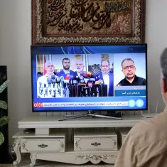 Un homme assis devant une télévision, qui affiche une conférence de presse avec plusieurs intervenants. La pièce est décorée d'une plante à côté de la télévision et d'un motif artistique sur le mur derrière.