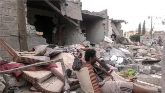 L'image montre une scène de destruction, probablement après un conflit ou une catastrophe naturelle. Au premier plan, un homme assis sur des matelas usés semble perdu dans ses pensées, entouré de débris. À l'arrière-plan, des bâtiments partiellement effondrés et des gravats jonchent le sol, témoignant des conséquences dévastatrices de l'événement. L'atmosphère est sombre et tragique, avec une sensation de désolation.