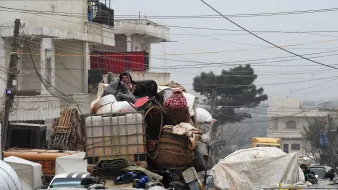 L'image montre une scène urbaine chargée, où des personnes sont perchées sur une pile de marchandises. Elles semblent se déplacer sur un véhicule surchargé, avec différents types de bagages et de matériaux. L'environnement est voilé par une lumière tamisée, peut-être due à un temps nuageux ou brumeux, et des câbles électriques traversent la scène. Les bâtiments en arrière-plan suggèrent un contexte urbain. Cette image évoque un sentiment de mouvement et de survie dans un espace urbain encombré.