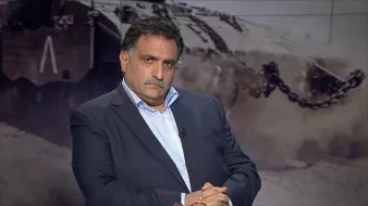 الصورة تظهر رجلًا يجلس بتعابير وجه جدية. يرتدي بدلة داكنة وقميص أبيض، ويداه متشابكتان أمامه. في الخلفية، يظهر مشهد غامض لمركبة عسكرية أو صورة حربية، مما يضيف جوًا من التوتر والدرامية للصورة. يبدو أن الرجل يتأمل أو يفكر بعمق.