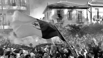 تظهر في الصورة حشود من الناس يحتفلون، مع مزيج من الدخان في الهواء. تتواجد مجموعة من المشجعين يحملون الأعلام واللافتات. يبدو أن هناك أجواء حماسية، مع تفاعل كبير بين المشاركين في الاحتفال. الخلفية تضم مباني سكنية، مما يضيف طابعاً حضرياً إلى المشهد.