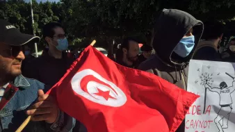 En la imagen se puede ver a un grupo de personas participando en una manifestación. Uno de los manifestantes sostiene una bandera roja con una estrella y una media luna blancas, símbolo nacional de Túnez. Otro individuo lleva un cartel que dice "Una idea no puede ser destruida", acompañado de una ilustración. La mayoría de los participantes utilizan mascarillas, y el ambiente parece ser de protesta pacífica. El fondo muestra árboles y un entorno urbano.