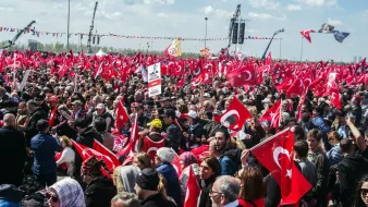 L'image montre une grande foule rassemblée en plein air, probablement lors d'un événement ou d'une manifestation en Turquie. De nombreuses personnes tiennent des drapeaux turcs, qui sont rouges avec un croissant blanc et une étoile. On peut voir des banderoles et des panneaux, et l'ambiance semble dynamique et engagée, avec un ciel bleu parsemé de nuages.