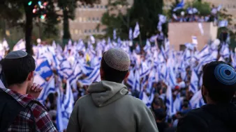 L'immagine mostra un gruppo di persone che osservano una grande manifestazione. Sullo sfondo si vedono moltissime bandiere israeliane sventolare tra la folla. Le persone nella parte anteriore dell'immagine sono viste di spalle; indossano kippah e sembrano concentrate sulla situazione davanti a loro. L'atmosfera sembra essere di impegno e partecipazione, con una grande presenza di persone che condividono una causa comune.
