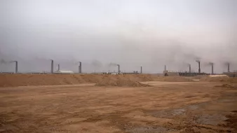 L'image montre un paysage industriel avec un sol désertique et nu. À l'arrière-plan, de nombreux cheminées émettent de la fumée dans une atmosphère brumeuse et chargée. Le ciel est grisâtre, ce qui donne une impression de pollution et de dégradation environnementale. Le terrain est principalement constitué de sable ou de terre, et l'ensemble dégage une atmosphère morose et industrialisée.