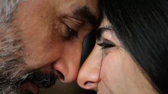 Cette image montre deux personnes, un homme et une femme, qui sont très proches l'un de l'autre. Ils touchent leurs fronts et semblent partager un moment intime. L'homme a une barbe grisonnante, tandis que la femme a des cheveux longs et foncés. Leur expression est douce : l'homme a un air serein, et la femme sourit joyeusement. L'ambiance de l'image évoque l'affection et la complicité entre les deux individus.