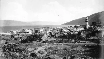 L'image montre un paysage historique d'un village côtier, probablement situé près d'un lac ou d'une mer, avec des bâtiments disposés le long de la promenade. Il y a un minaret ou une tour surmontée d'un dôme, indiquant la présence d'une culture musulmane. En arrière-plan, on peut voir des collines qui encadrent le paysage. Le ciel est légèrement nuageux, créant une ambiance tranquille. L'ensemble de l'image a un aspect noir et blanc, ce qui lui donne un caractère ancien.