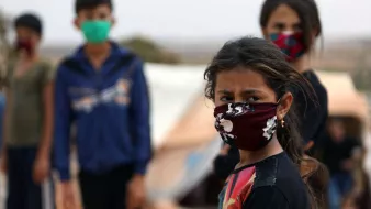 L'image montre un groupe d'enfants, dont une fille au premier plan, portant des masques. La jeune fille a les cheveux bruns et des yeux expressifs, semblant préoccupée. En arrière-plan, d'autres enfants se tiennent, également masqués, dans un environnement qui semble être un camp. L'atmosphère semble chargée d'émotion et de défi. Les couleurs des masques varient, ajoutant une touche de diversité visuelle à la scène.