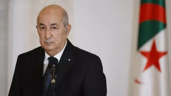 L'image montre un homme en costume sombre, regardant vers l'avant avec une expression sérieuse. Il se tient devant un microphone, et en arrière-plan, on peut voir un drapeau. Le drapeau présente des couleurs vertes et rouges avec une étoile et un croissant. L'ambiance de l'image semble solennelle, suggérant un contexte formel ou officiel.