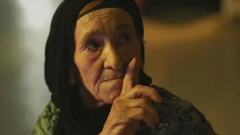 L'image montre une femme âgée avec un regard pensif. Elle porte un foulard noir qui encadre son visage. Son expression est sérieuse et elle semble faire un geste de silence avec son doigt posé sur ses lèvres. La lumière dans le fond est douce, créant une ambiance calme et introspective. Les détails de son visage reflètent une certaine sagesse et vécu.