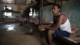 L'image montre un espace intérieur qui semble être une chambre ou un dortoir. On y voit plusieurs lits superposés et des personnes assises ou allongées. Un jeune homme est assis sur le bord d'un lit, regardant pensivement au loin. Il porte un débardeur et un short, et ses pieds sont nus. Les murs de la pièce sont nus et usés, avec des traces d'humidité, ce qui indique un environnement modeste et probablement surpeuplé. La lumière naturelle pénètre par une fenêtre, ajoutant une certaine ambiance à la scène.