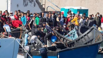 L'image montre un groupe de personnes sur des embarcations, probablement dans un port ou une zone côtière. Les individus semblent rassemblés autour de plusieurs bateaux, avec une diversité de vêtements, certains portant des masques. L'environnement est urbain, et il y a des structures en béton visibles en arrière-plan. On peut noter une atmosphère de mouvement et d'activité parmi les personnes présentes sur les bateaux.