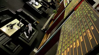 The image depicts a financial trading environment, likely within a stock exchange. In the foreground, there is a large electronic display board showing various financial data, such as stock prices and indices, with green and red text indicating changes in value. Below the board, there are several black leather seating areas where traders or visitors might be sitting. One individual, dressed in traditional attire, appears to be engaged with a computer or taking notes. The overall atmosphere suggests a busy and professional financial setting.