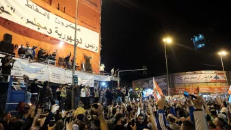 The image captures a large crowd gathered in an urban area, likely during a rally or protest. People are raising their hands, some holding flags, and others are holding up their phones, possibly recording the event. In the background, there is a large banner that appears to convey a political message, and a group of speakers is visible on a stage or elevated platform. The atmosphere seems energetic and filled with a sense of purpose, with bright lights illuminating the scene at night.