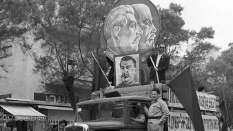 تصویر نشان‌دهنده یک خودروی قدیمی است که در یک مراسم راهپیمایی یا تظاهرات شرکت کرده است. بر روی ماشین بزرگ‌نمایی از چهره‌های افرادی با موهای کوتاه و مقدار زیادی نماد و پرچم‌هایی وجود دارد. فردی در حالت ایستاده بر روی خودرو قرار دارد و به نظر می‌رسد که در حال صحبت یا اشاره به تماشاگران است. پس‌زمینه شامل گروهی از مردم است که در کنار خیابان ایستاده‌اند، و فضایی از شور و اشتیاق را در این رویداد به تصویر می‌کشد.