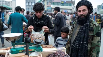 The image depicts a bustling market scene, likely in Afghanistan. In the foreground, there is a vendor displaying various types of dates in baskets and boxes. The vendor, dressed in a traditional outfit and a black turban, stands next to a scale. Another young man is shown packing dates into a bag, while a young boy looks on. The background features a crowd of people, creating a lively atmosphere, with buildings visible in the distance. The overall scene reflects daily life and commerce in a market setting.
