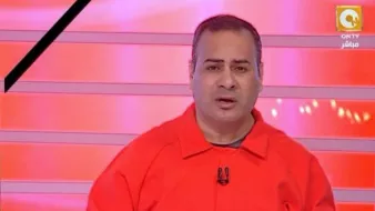L'image montre un homme en train de parler devant un fond rouge avec des lignes lumineuses. Il porte une combinaison orange et semble être en train de faire une présentation ou de commenter quelque chose. Son expression faciale est sérieuse et il tient un stylo dans sa main. Le décor et son apparence suggèrent qu'il s'agit d'un animateur ou d'un commentateur à la télévision.