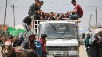 L'immagine mostra un gruppo di persone che si trovano su un camion, alcuni di loro sono seduti sul retro del veicolo, mentre altri sono in piedi. Sul lato destro ci sono persone su un carro trainato da un animale, che sembra essere un cavallo. In fondo si intravedono tende e strutture, suggerendo un contesto urbano o di accampamento. L'atmosfera sembra vivace, con diverse persone che interagiscono tra loro.