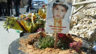 L'immagine mostra un tributo commemorativo collocato su un terreno. Al centro, c'è un cartello che presenta un ritratto di un bambino, adornato con un abbigliamento tradizionale. Attorno al cartello ci sono fiori freschi e piante, posizionati in una gomma di automobile. Il tutto è circondato da un ambiente duro, con rocce visibili e alcune persone in fondo che sembrano partecipare a una commemorazione. La data sul cartello indica un giorno significativo, possivelmente legato alla memoria del bambino.